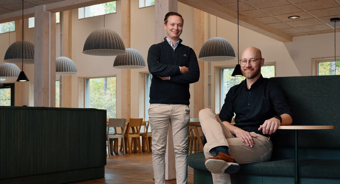 Medarbetaren Gabriel står upp och kollar in mot kameran, kollegan Daniel sitter i en soffa bredvid och kollar in mot kameran.